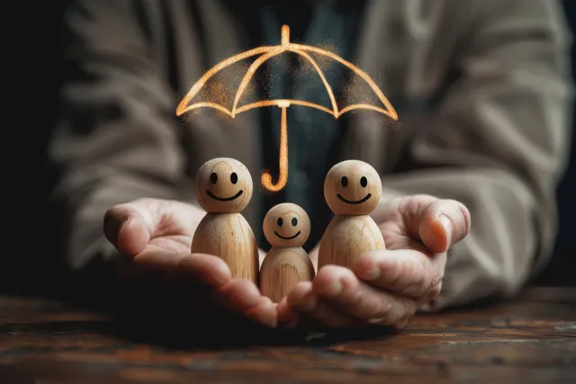 A businessman’s hands hold smiling wooden family figures under an open umbrella symbolizing insurance protection.