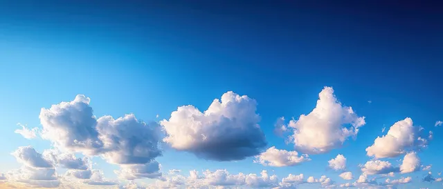 Fluffy white cumulus clouds float across a vivid blue sky above a bright sunny horizon.