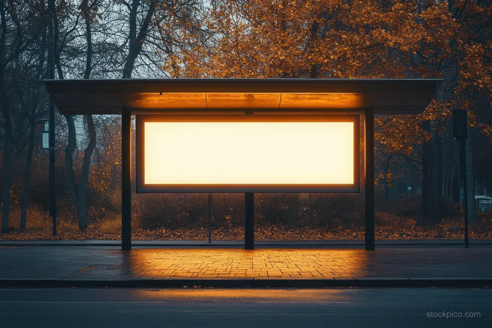 Empty bus stop shelter with a blank glowing billboard panel at dusk in an autumn street scene.