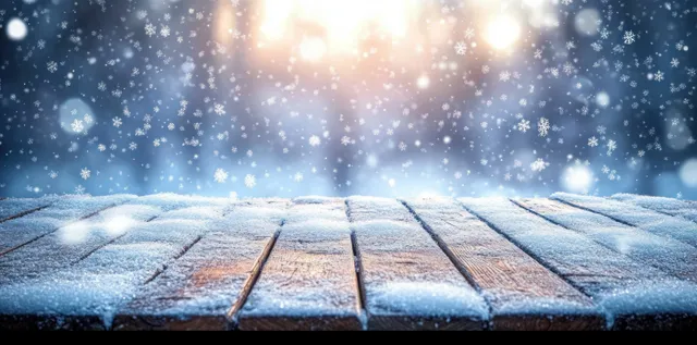 Snow covers a wooden deck while glowing lights blur in the blue background as snowflakes fall.