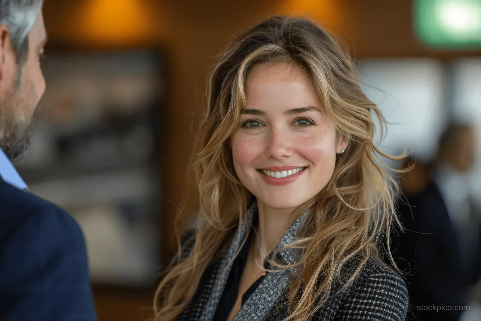 Smiling businesswoman shakes hands with a male executive in an office while others blur in the background.