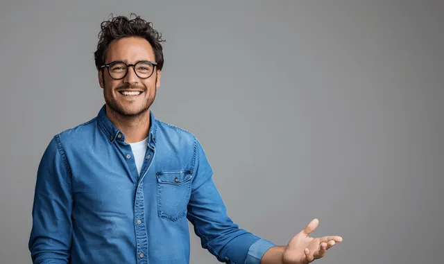 Smiling man in a blue shirt extends an open palm gesture on a plain gray background.