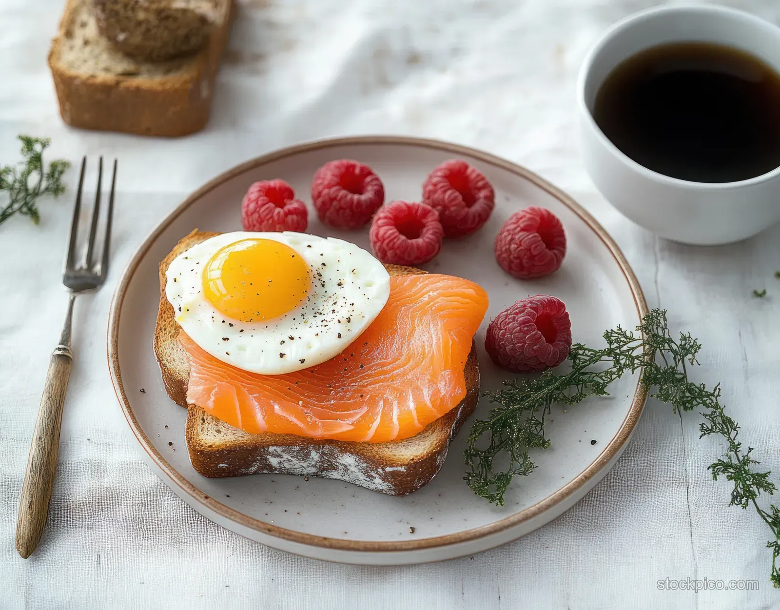 Toasted bread topped with smoked salmon and a sunny-side-up egg sits on a plate next to raspberries and black coffee.