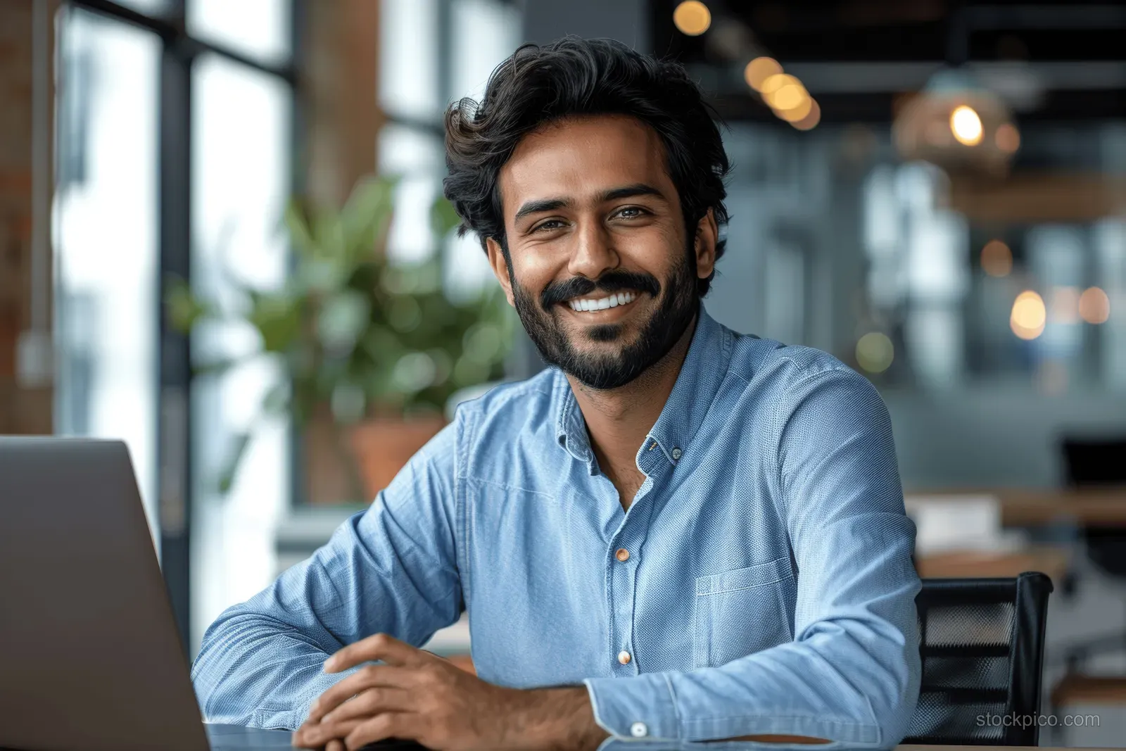 Smiling Indian businessman works on a laptop at a desk in a modern office with warm blurred lights.