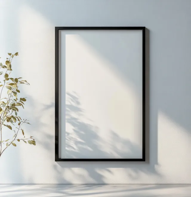 A black framed blank art print is mounted on a light wall with soft sunlight shadows.