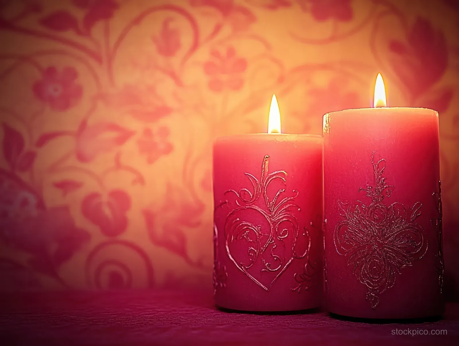 Two lit red decorative candles with ornate embossed patterns glow on a textured surface beside a warm floral background.