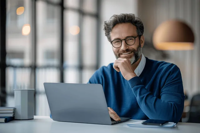 A man in a blue sweater works on a laptop at a well-lit office desk with a pen under his chin.