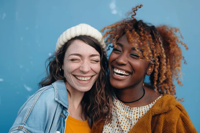 Two women best friends laugh and smile closely together in front of a solid blue studio background.