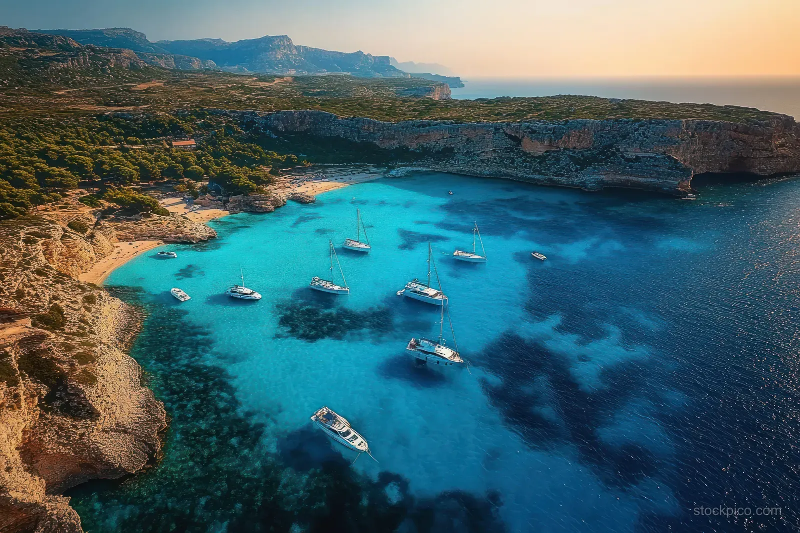 Aerial view shows sailboats anchored in a crystal-blue bay near rugged green cliffs and distant rocky mountains.