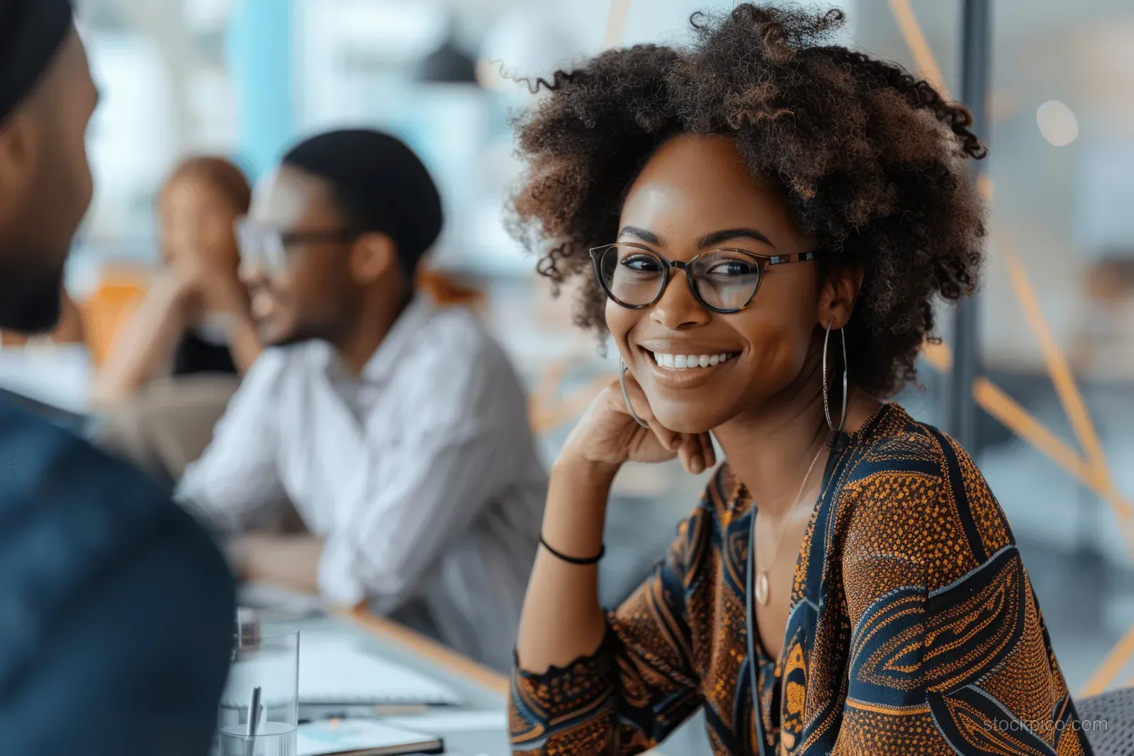A smiling Black businesswoman in glasses leads a boardroom meeting while colleagues discuss in the background.