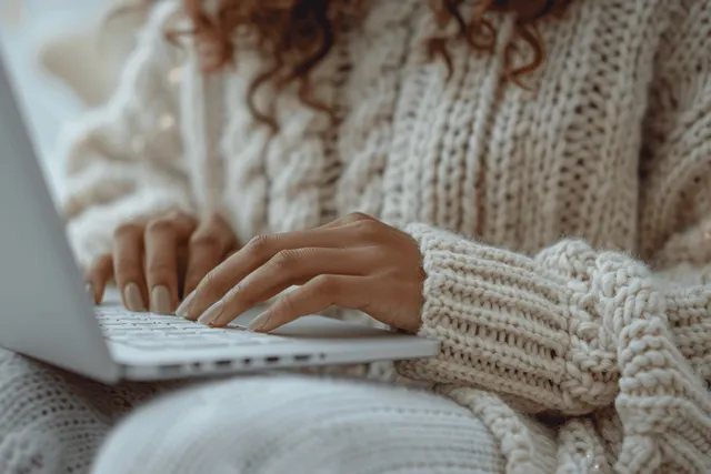 Woman typing on laptop keyboard while working online from cozy beige sweater desk