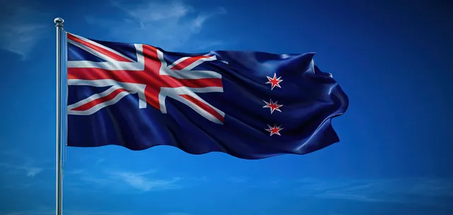 New Zealand flag with Union Jack and stars waving on a pole against a bright blue sky.