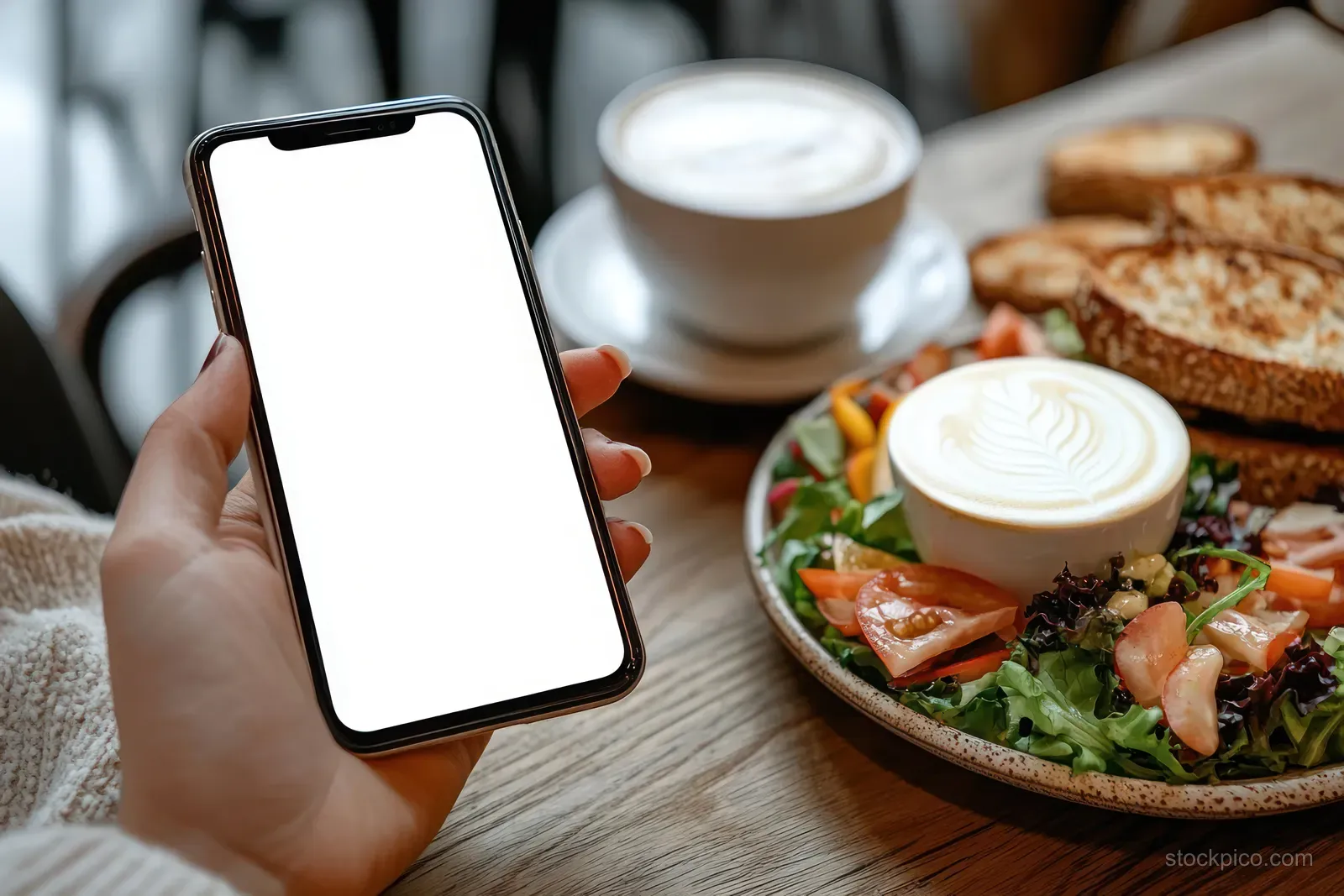 A hand holds a smartphone with a blank screen next to salad, coffee cups, and toasted bread on a wooden table.