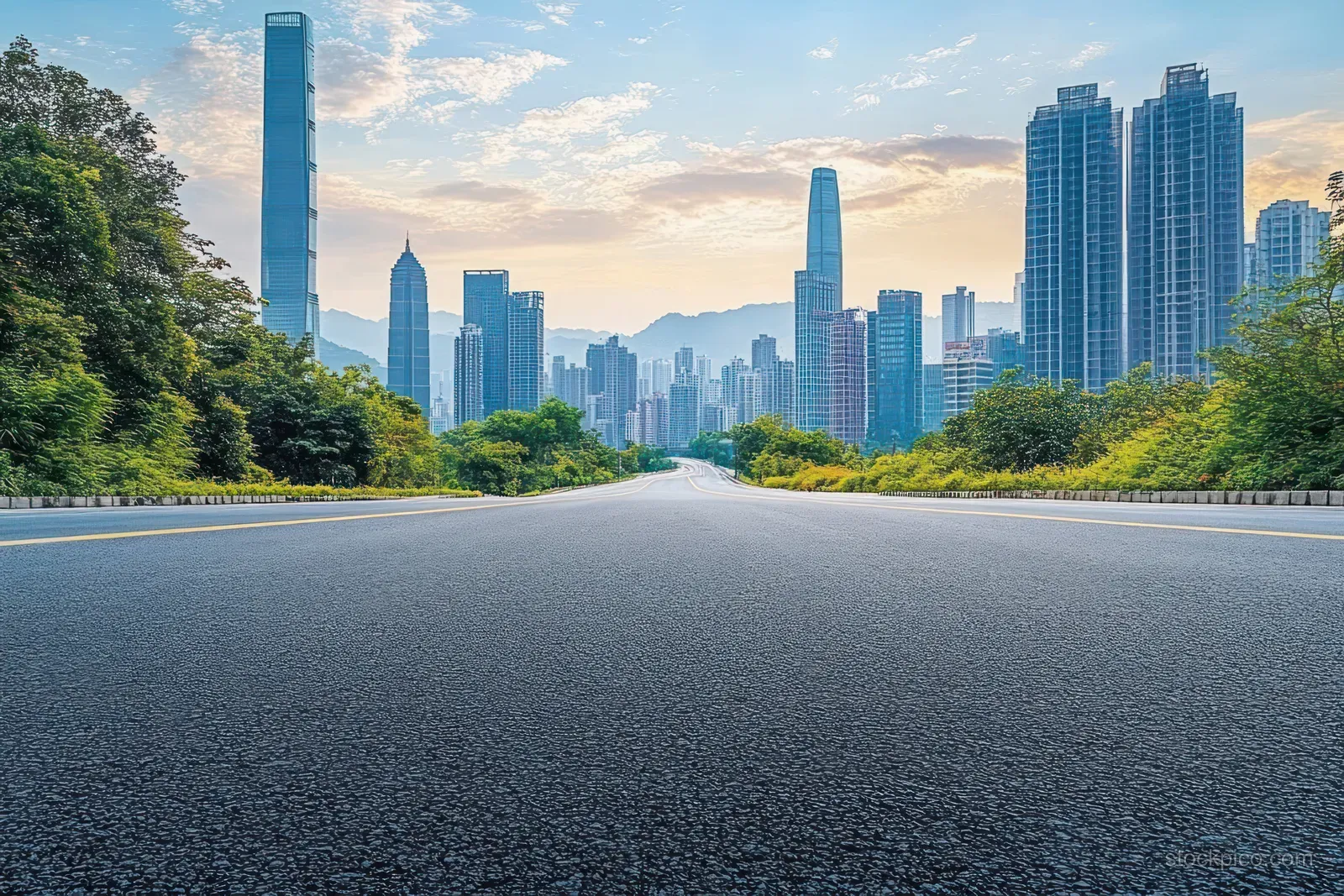 Empty asphalt road vanishing toward a modern skyscraper skyline with green trees under sunset clouds.