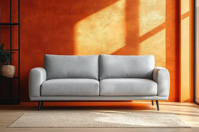 Grey modern sofa against a textured orange wall with geometric sunlight shadows on the wall and light wooden floor.