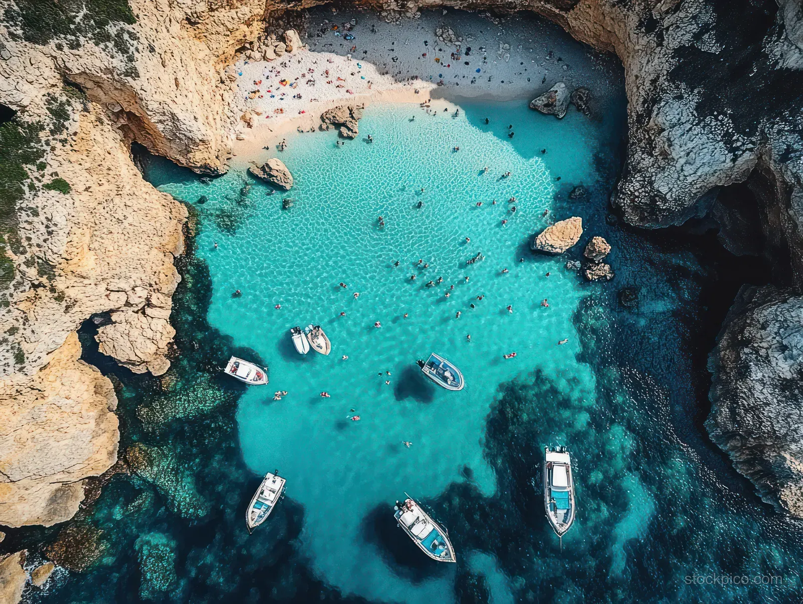 Aerial view shows turquoise water in a rocky cove with anchored boats and swimmers near a sandy beach.