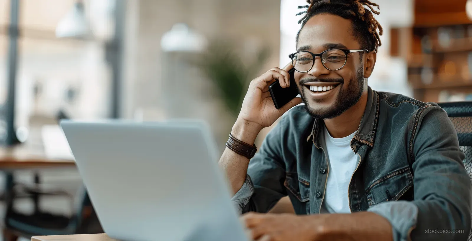 A young businessman wearing glasses speaks on a smartphone while typing on an open laptop in a modern office.