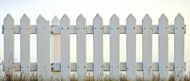 Weathered white picket fence with pointed tops stands behind dry grass beneath a clear blue sky.