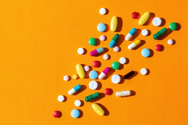 Colorful pills and capsules scattered on a bright orange background in a flat-lay composition.