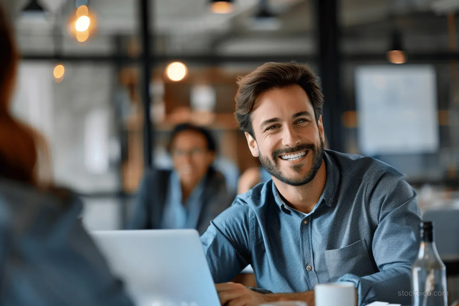 Smiling man at a laptop table with coworkers collaborating in a modern office background.