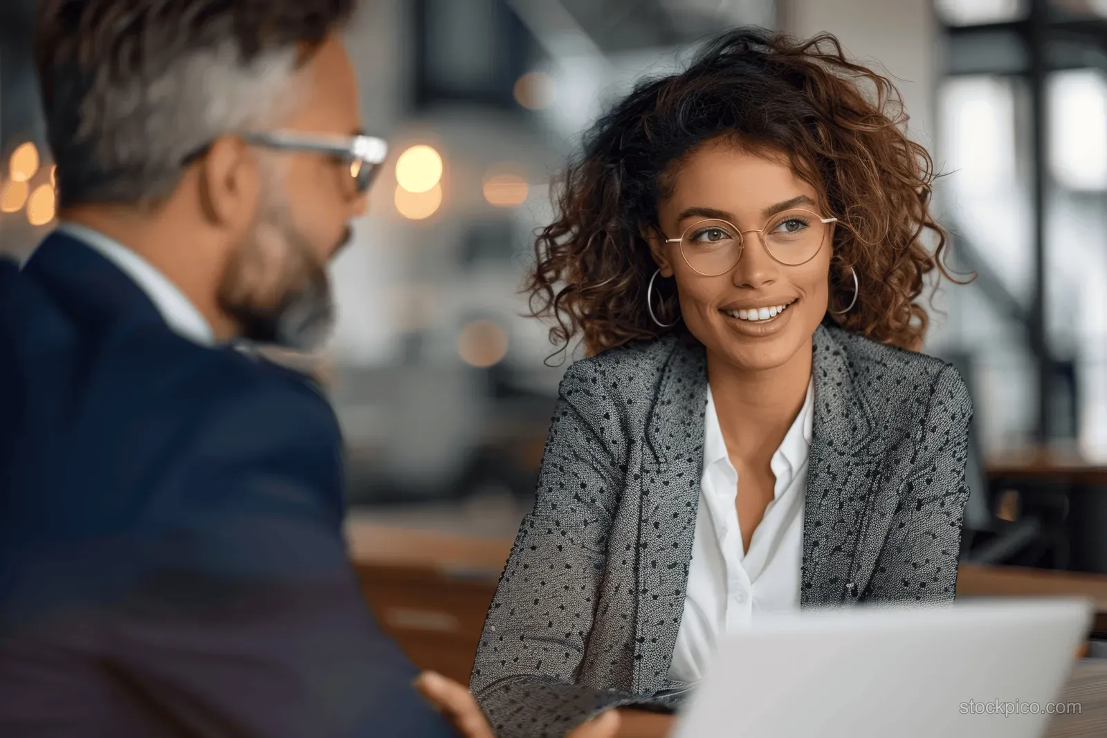 Curly-haired businesswoman smiles while reviewing a laptop with a colleague during an office meeting.