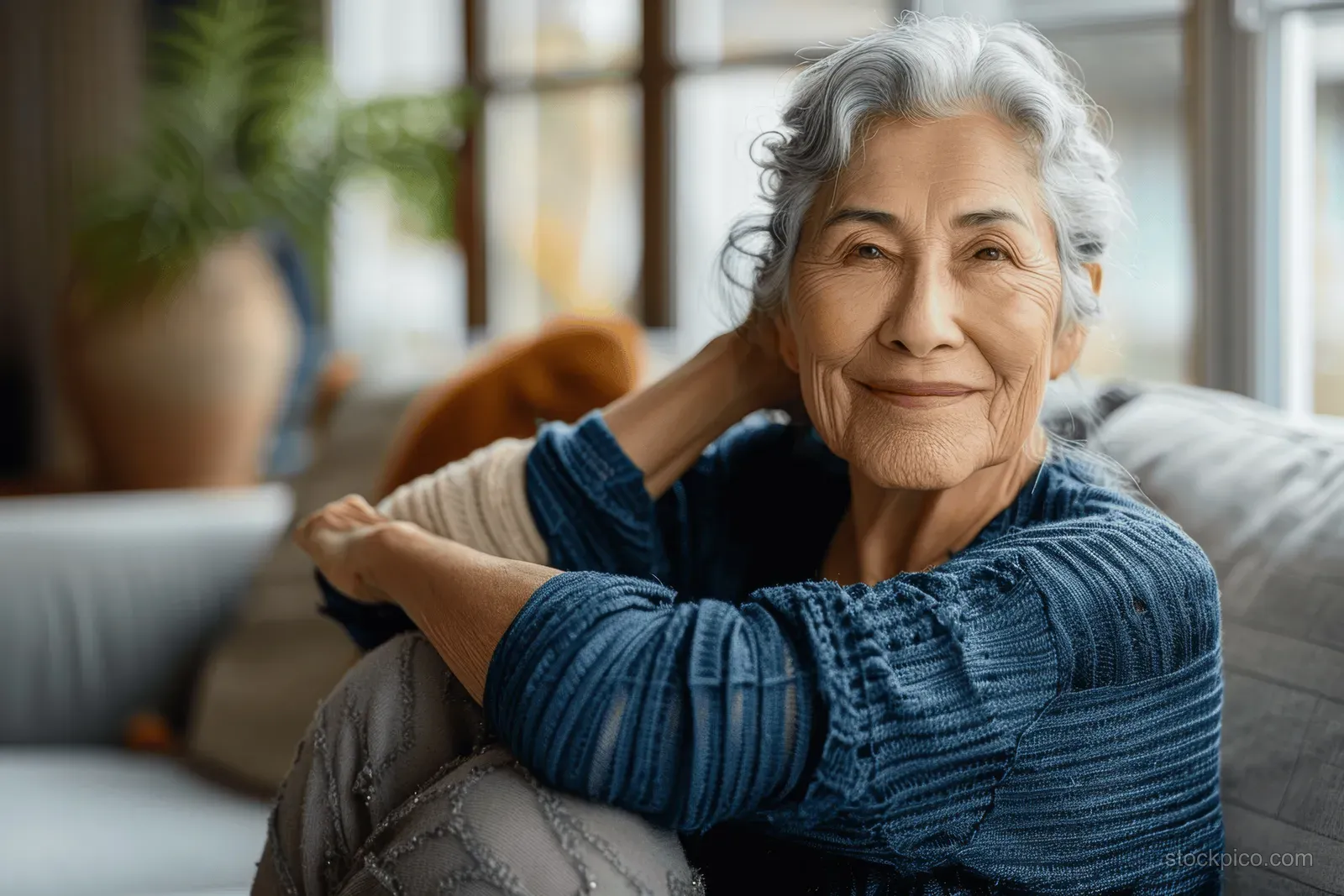 A senior woman stretches her arms while seated on a sofa in a bright living room with large windows.
