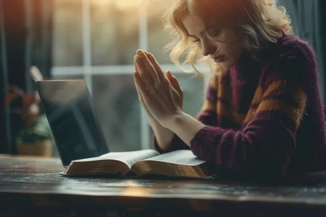 Woman prays beside an open Bible and laptop on a wooden table in warm window light.
