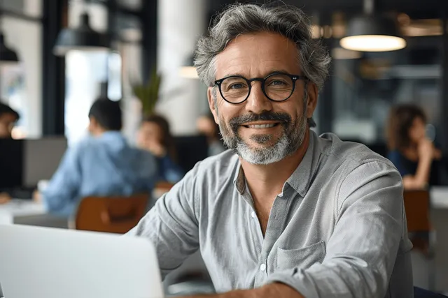 Smiling mature businessman teacher with glasses sits at desk using laptop in modern office
