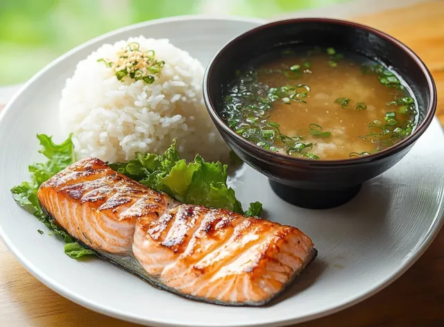 Grilled salmon fillet served with white rice and miso soup garnished with chopped green onions on a white plate.