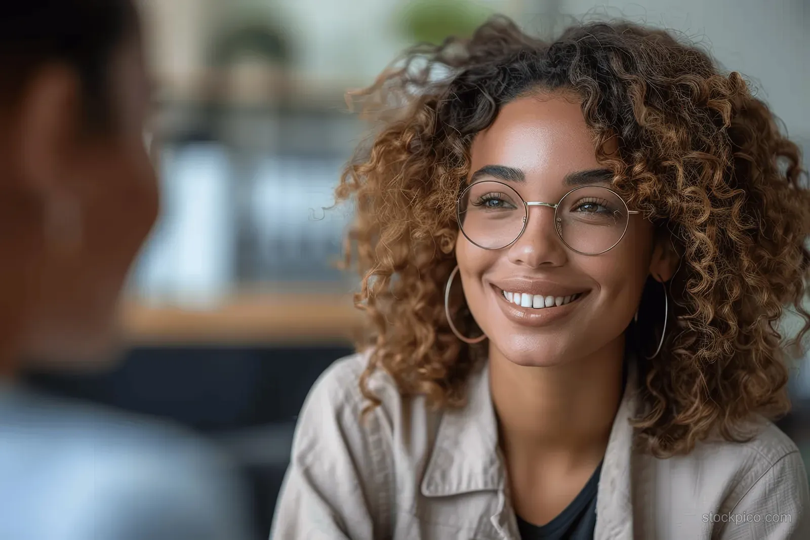 Curly-haired therapist with glasses smiles during a one-on-one counseling conversation in an office.