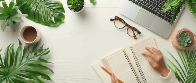 Student’s hands type on a laptop while an open notebook and glasses sit on a white desk with plants.