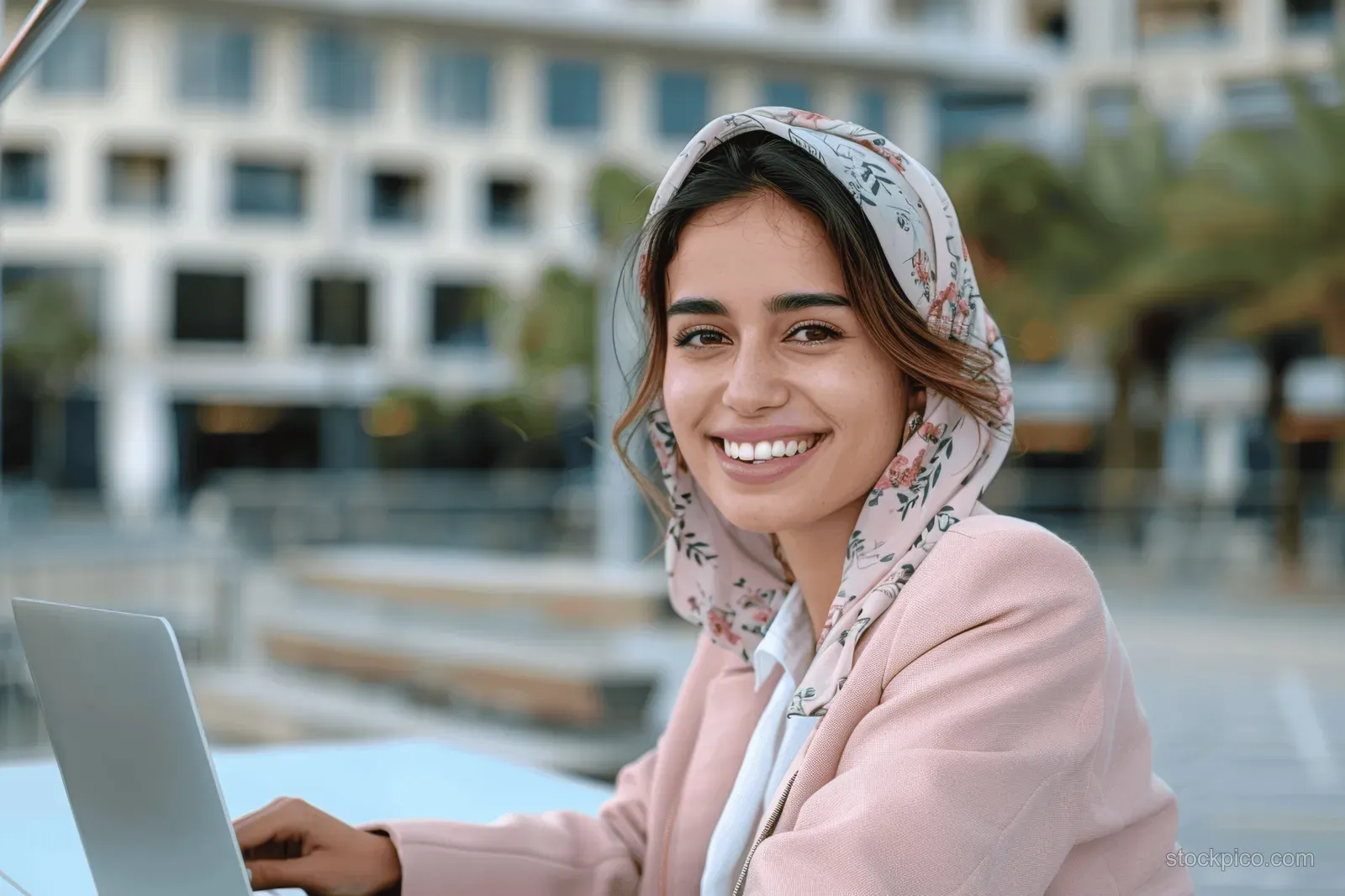 Smiling young businesswoman in a pink jacket works on a laptop outdoors near modern building architecture.