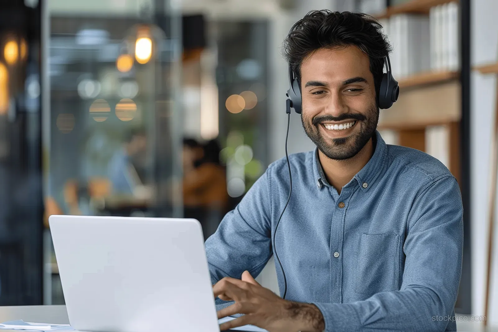 Smiling Indian businessman with a headset uses a laptop for a video call in a modern glass-walled office.