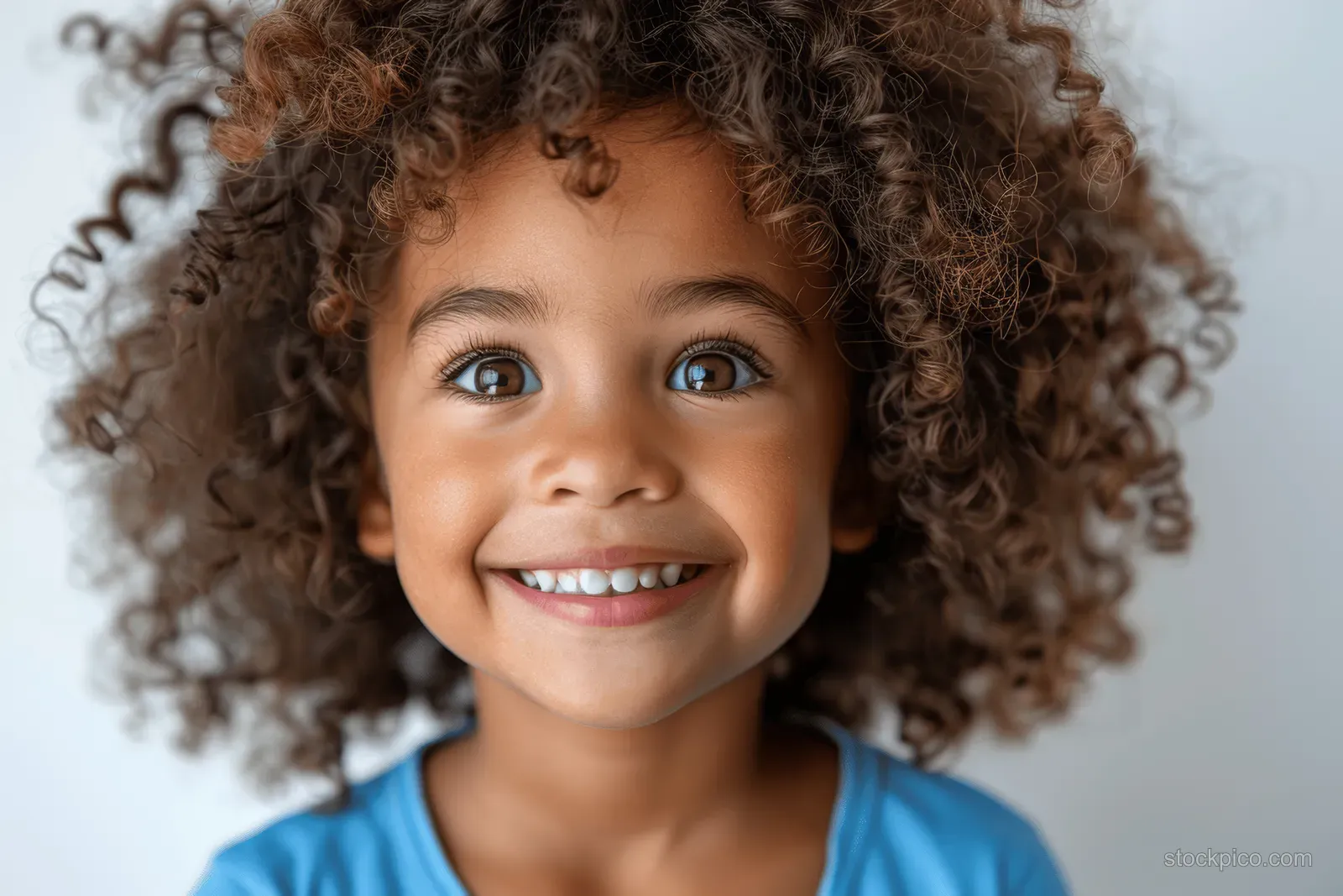 A smiling child with curly hair wearing a blue shirt, photographed against a white background.