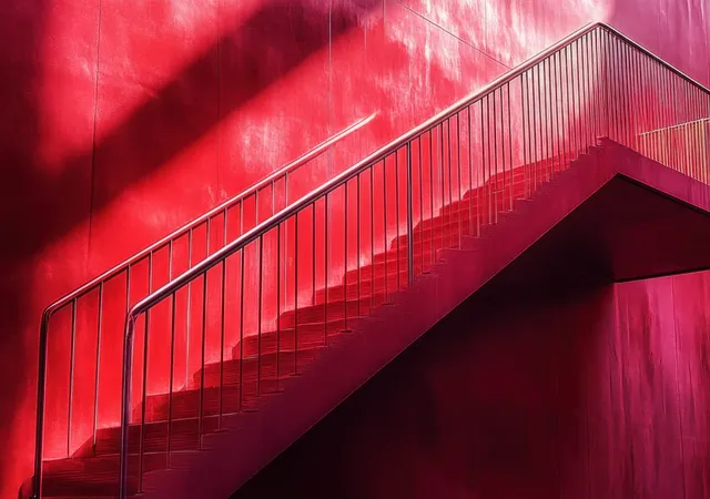 A red staircase with metal railing casts sharp geometric shadows on a vivid red wall in bright sunlight.