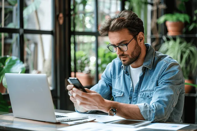 Adult entrepreneur in glasses uses a smartphone while reviewing paperwork next to an open laptop at an office desk.