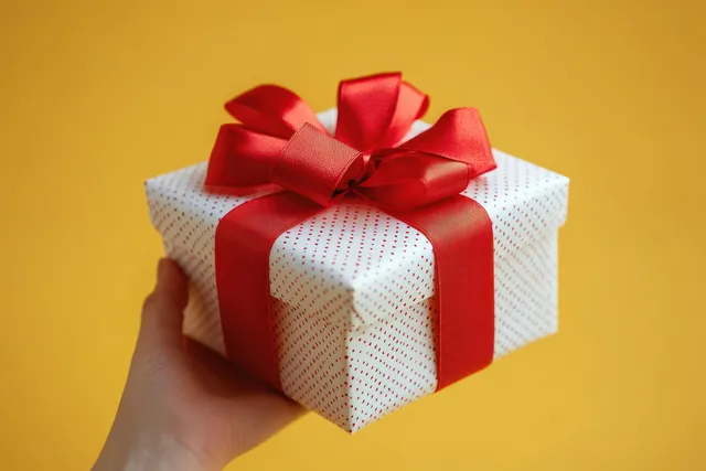 A hand holds a white gift box wrapped with red dots and tied with a shiny red ribbon bow on a bright yellow background.
