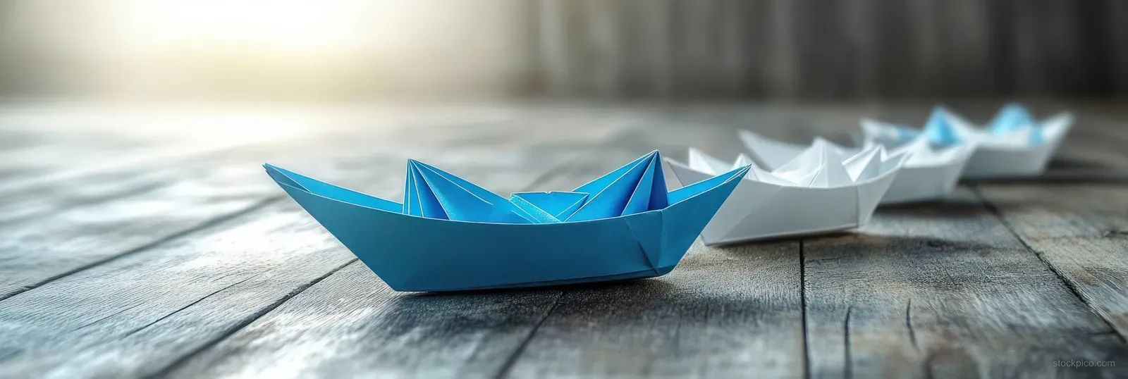A blue paper boat leads white origami boats along a weathered wooden surface with soft shadows.