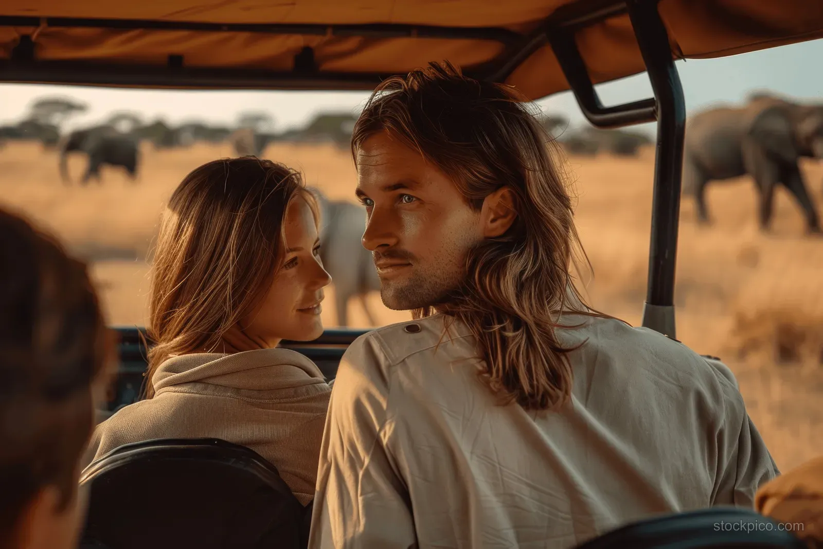 A young couple rides an open safari jeep while elephants walk across the savanna outside.