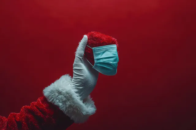 A red-coated Santa gloved hand holds a teal surgical face mask against a deep red backdrop.