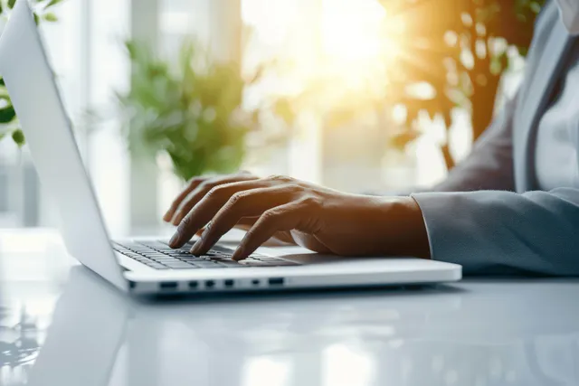 Hands typing on a laptop at a desk with warm sunlight streaming through office windows.