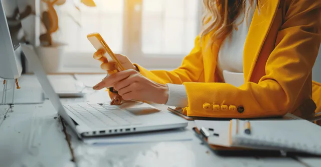 Businesswoman in a yellow blazer points at her smartphone screen while typing on a laptop at an office desk.