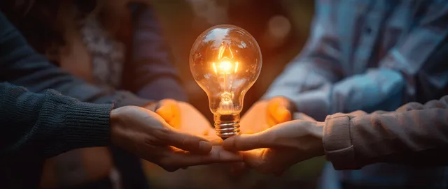 Several hands hold a glowing light bulb together in a dark setting.