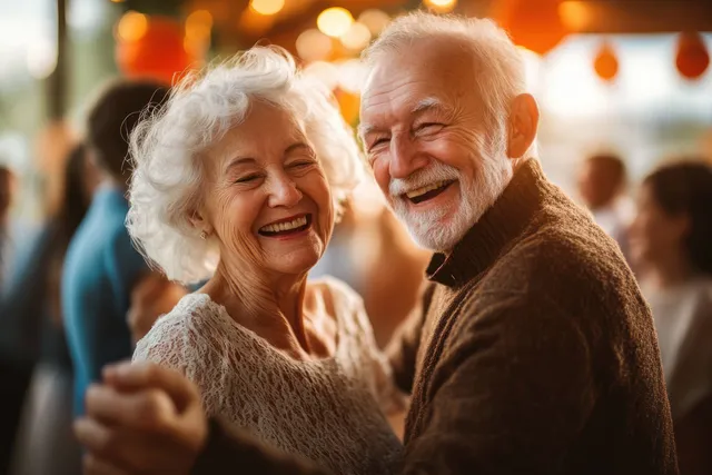 An elderly couple dances closely and smiles at a warmly lit indoor social event with blurred guests behind them.