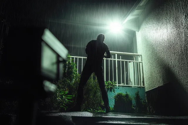 A masked burglar climbs a house wall at night as a surveillance camera spotlight casts sharp shadows.