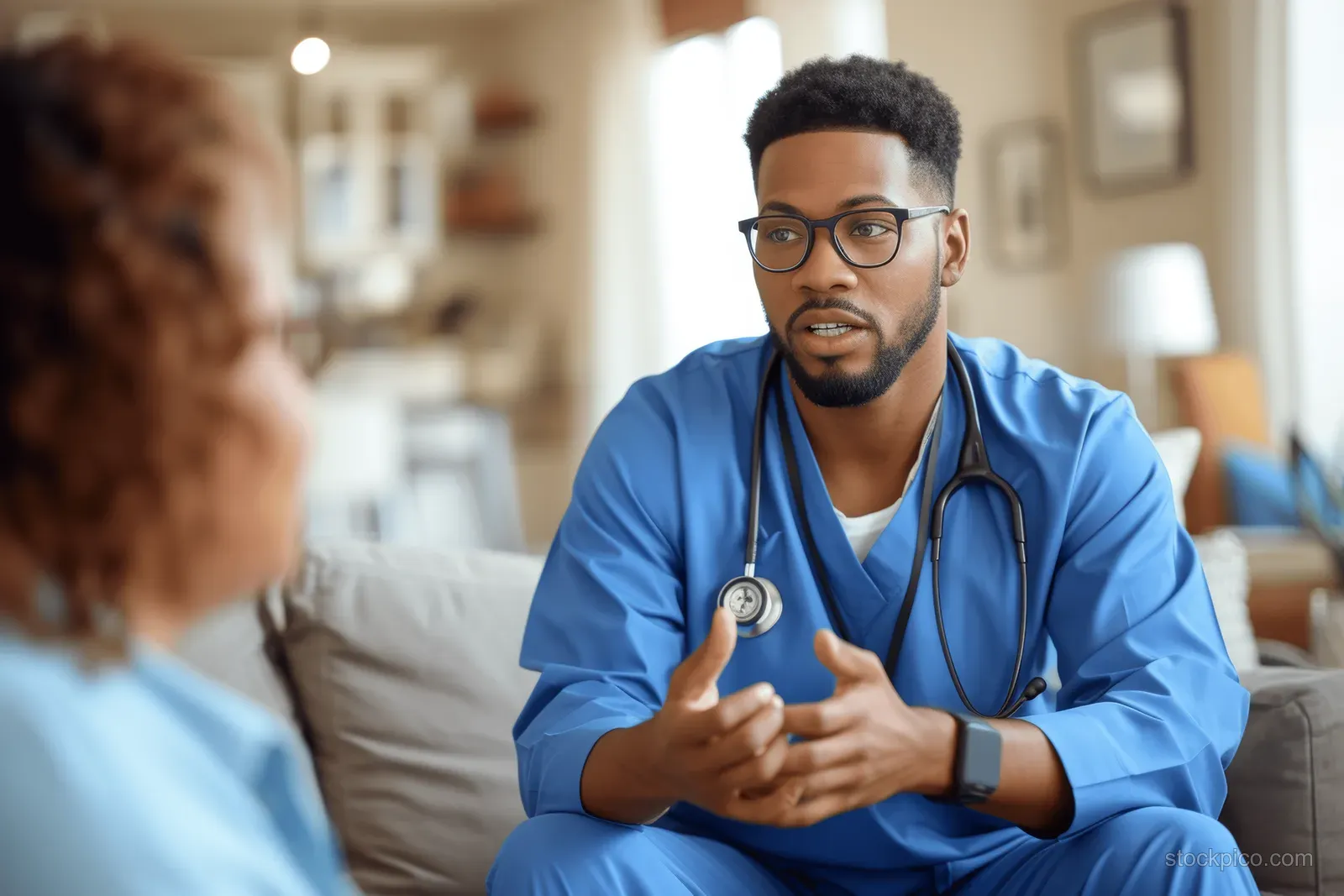 A doctor in blue scrubs speaks with a senior patient in a living room during a home healthcare visit.