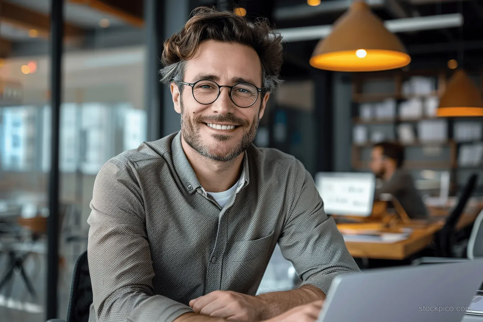 Man in glasses smiling at a laptop on a desk in a modern office with blurred background coworkers.