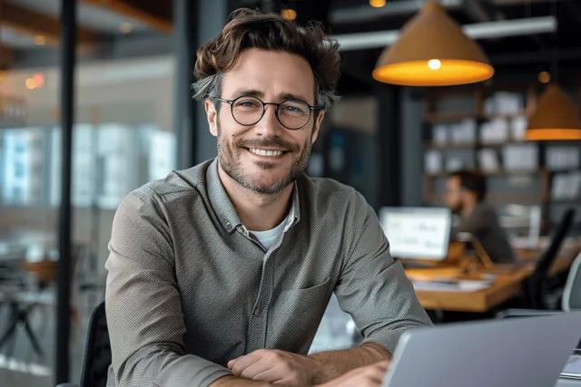 Man in glasses smiling at a laptop on a desk in a modern office with blurred background coworkers.