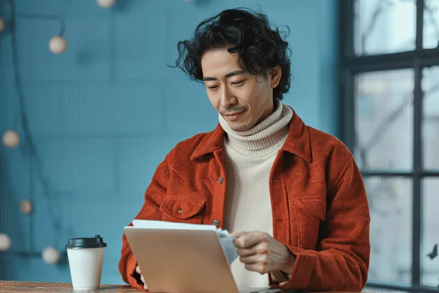 Asian businessman reviewing documents on laptop desk in natural light office window