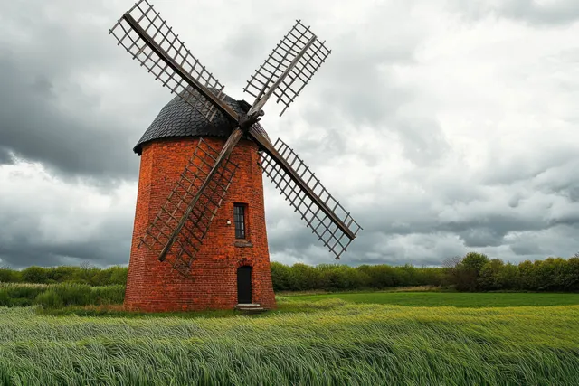 An old brick windmill with wooden blades stands in a green field under a cloudy gray sky.